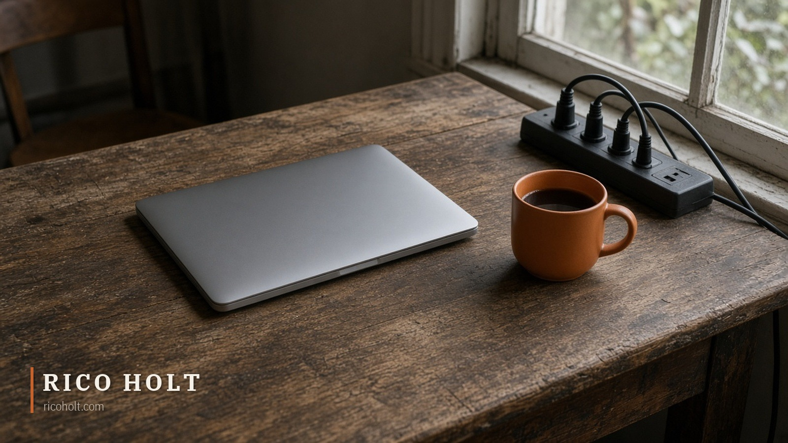 A worn timber desk by a window in soft morning light with a closed grey laptop, a full black power board and one ochre ceramic mug