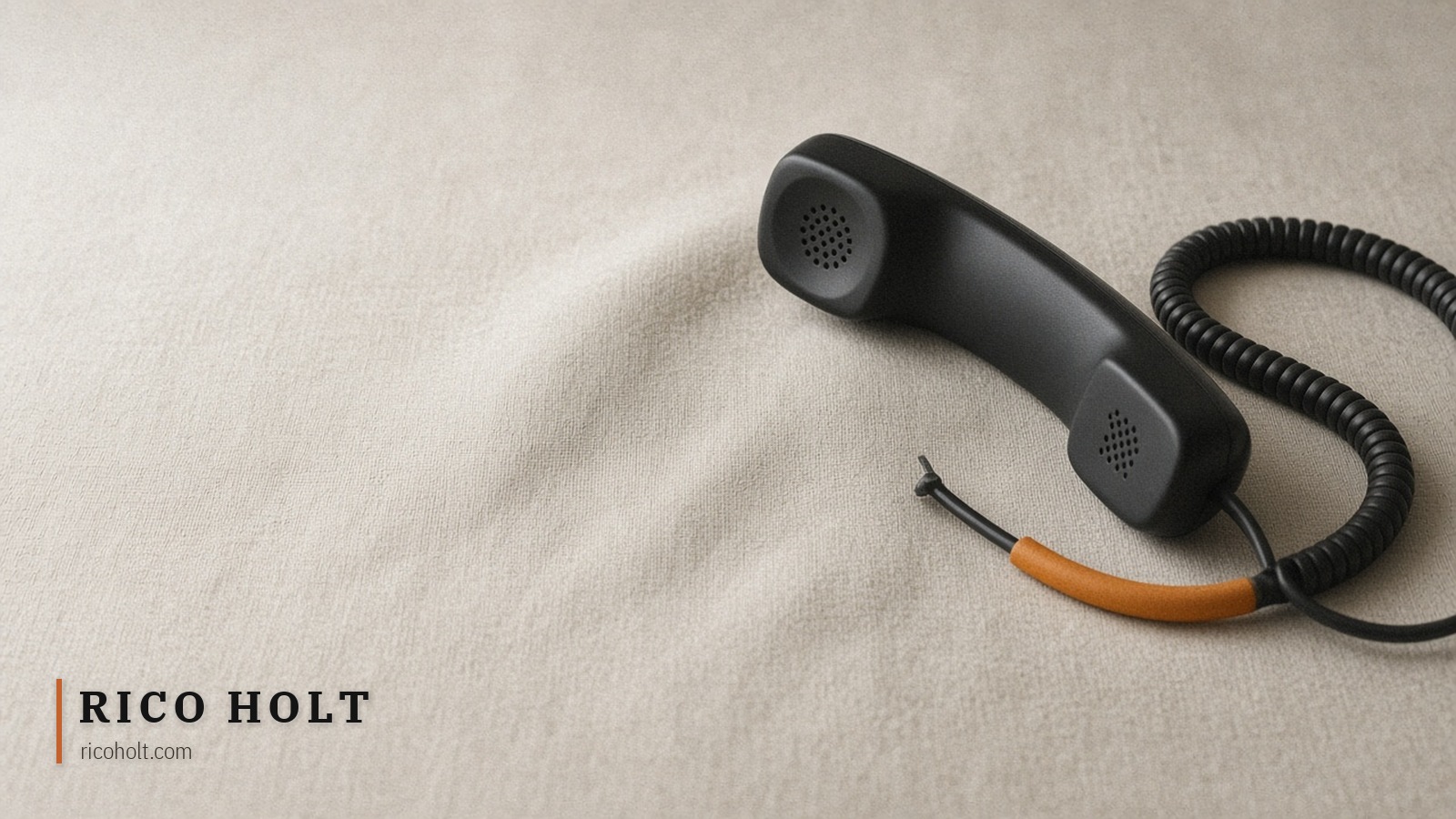 Plain black landline handset off the hook on pale warm-grey linen, coiled cord with one short matte ochre segment