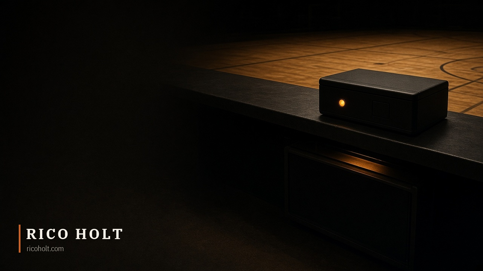 A matte-black shot clock controller with a glowing amber indicator lamp on a dark scorer's table ledge overlooking a spotlit hardwood basketball court at night