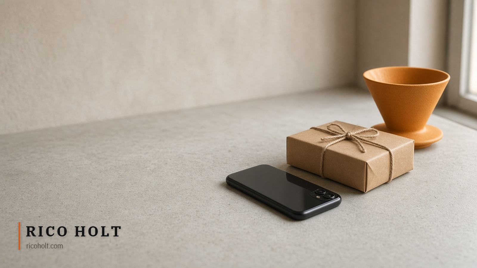 Pale concrete cafe counter in soft window light with a face-down black phone, a string-tied kraft parcel and one ochre ceramic coffee dripper