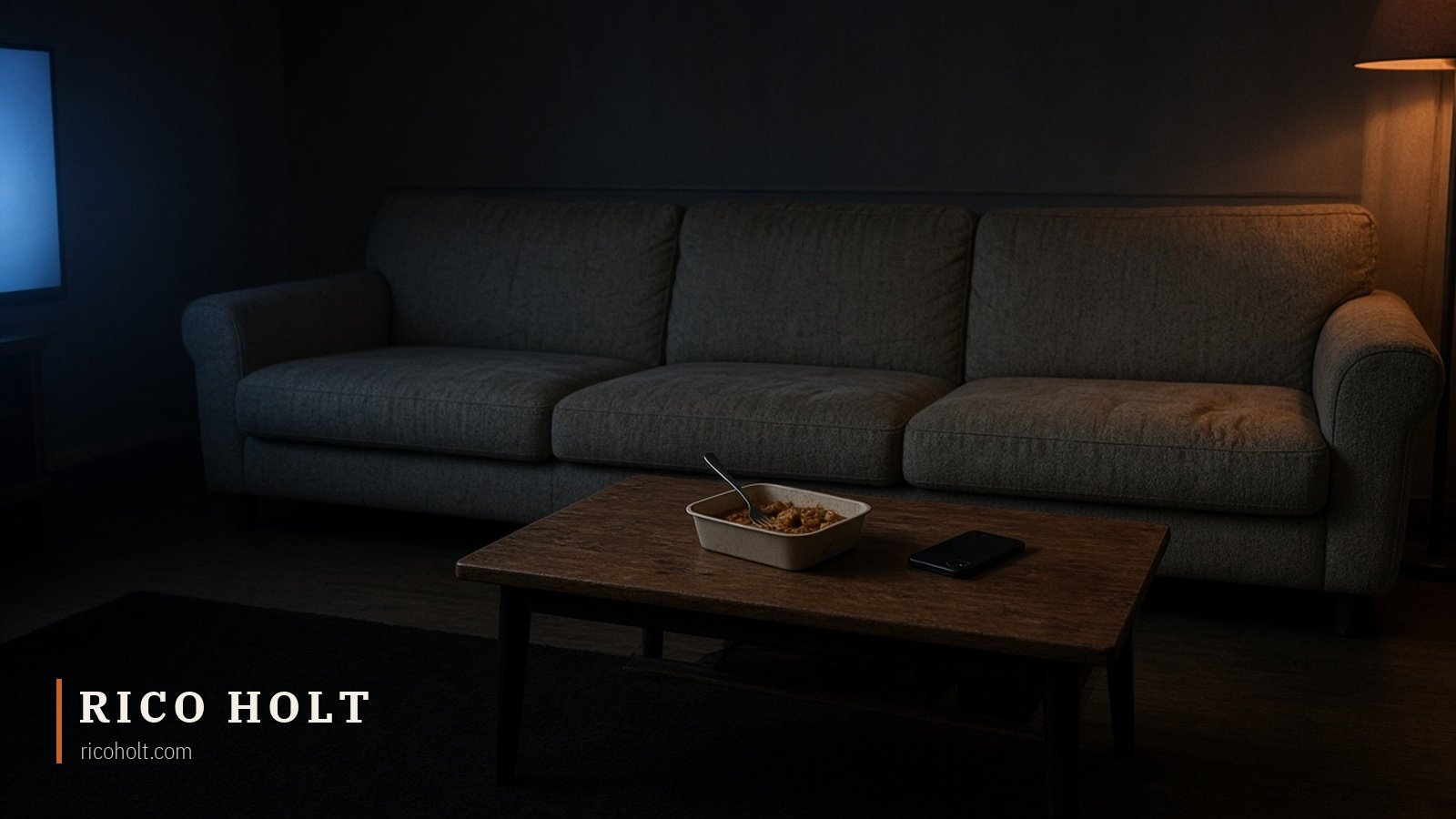 A dim suburban lounge room at night: an empty grey three-seat couch, a timber coffee table holding one takeaway curry container with a fork in it and a phone lying face down, warm amber floor lamp glow on the right and cold blank TV light from the left